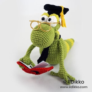 Op de afbeelding: Een groen gehaakt hagedisfiguur met een zwarte afstudeerhoed met een gele kwast, een goudkleurige bril en een zwarte das. De hagedis houdt een open rood boek met witte pagina's vast. De tekst "Ildikko www.ildikko.com" is zichtbaar.