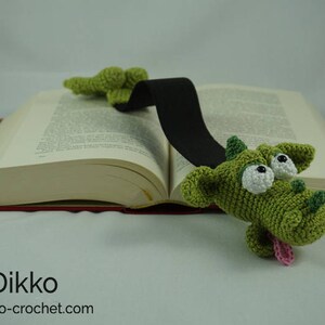 May include: A green crocheted dragon bookmark with large white eyes and a pink tongue. The bookmark is attached to a black ribbon and is placed on a book.