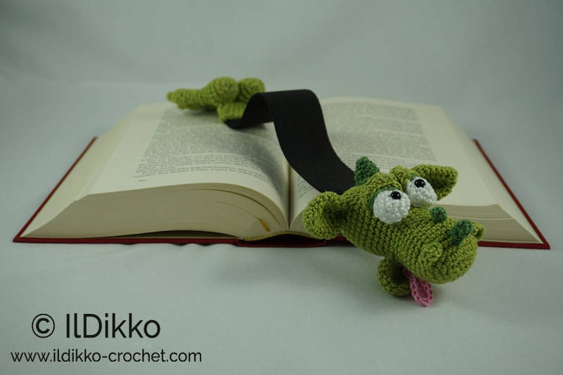 May include: A green crocheted dragon bookmark with large white eyes and a pink tongue. The bookmark is attached to a black ribbon and is placed on a book.
