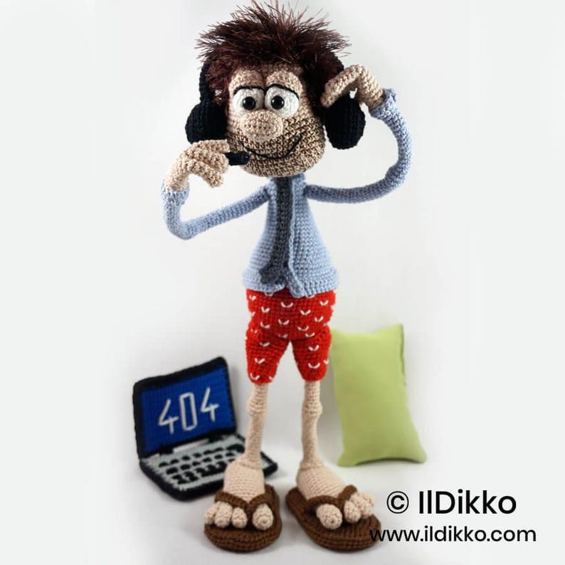 May include: A crocheted doll with brown hair, wearing a light blue shirt, red shorts with white patterns, and brown sandals. The doll wears black headphones with a microphone. Beside the doll is a small crocheted laptop displaying 404 text.