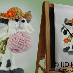 May include: A crocheted cow wearing a brown and orange hat with a flower. The cow is holding a green stem with an orange flower. The cow is standing on a white surface.