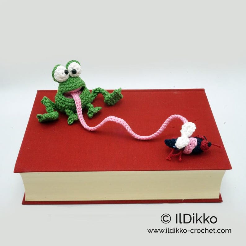 May include: A green crocheted frog with a pink tongue catching a red and white crocheted fly. The frog and fly are on top of a red book with the text "&copy; Ildikko www.ildikko-crochet.com".