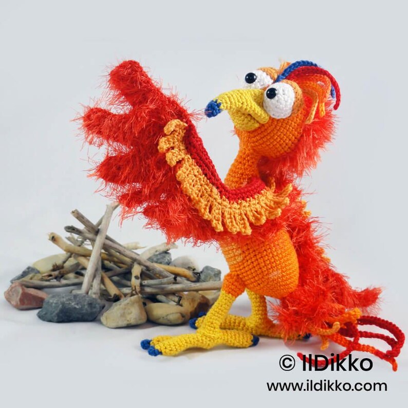 May include: A crocheted phoenix figurine featuring a bright orange body, yellow legs, and vibrant red feathered wings made of textured yarn. The creature has large white eyes with black pupils, a yellow beak, and blue accents on the head and feet.