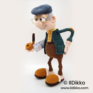 May include: A handmade crochet doll of an elderly man wearing a blue cap, glasses, and a green jacket over a yellow shirt. The doll holds a pipe and a booklet. The doll has brown pants and large, orange shoes with white trim. The doll is a whimsical, handcrafted item.