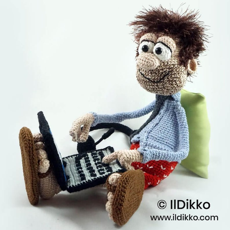 May include: A crocheted doll with messy brown hair, large eyes, and a wide smile, wearing a light blue shirt and red patterned shorts. The doll sits with brown shoes, using a black and gray crocheted laptop with a black headset attached.