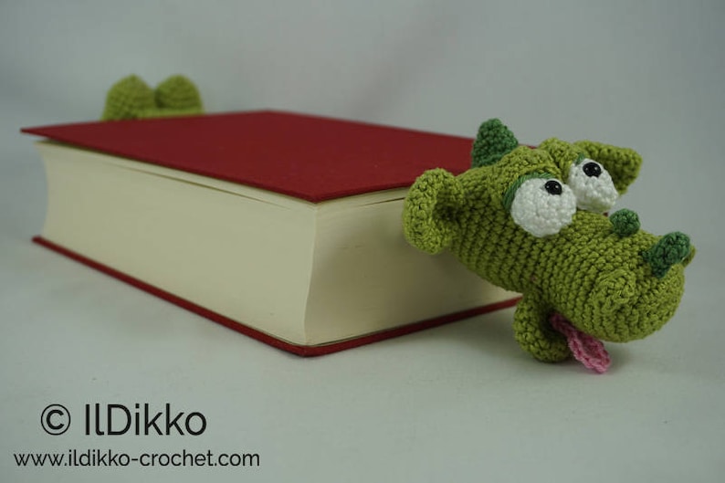 May include: A green crocheted dragon bookmark with white eyes and a pink tongue, holding a red book closed.