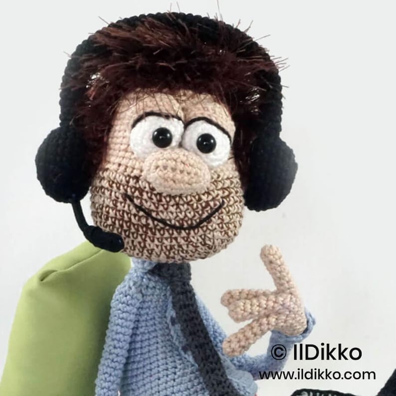 May include: A crocheted doll featuring a light blue shirt, a dark tie, and brown hair. The doll wears black headphones with a microphone attachment. The face has large white eyes with black pupils and a stitched smile. Text reads IlDikko ildikko.com.