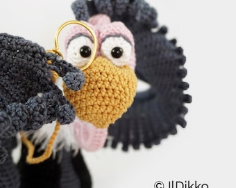 May include: A crocheted vulture figurine featuring dark gray wings, a light pink head, a textured mustard yellow beak, and large white eyes with black pupils. A gold-colored metal ring is attached to the wing. Text reads: © ILDikko www.ildikko-crochet.com