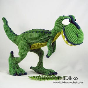 Può includere: Giocattolo di dinosauro verde all'uncinetto con un ghigno dentato, in piedi su una superficie bianca con una foglia verde in primo piano. Il dinosauro ha una pancia gialla e accenti blu sui suoi piedi e occhi.