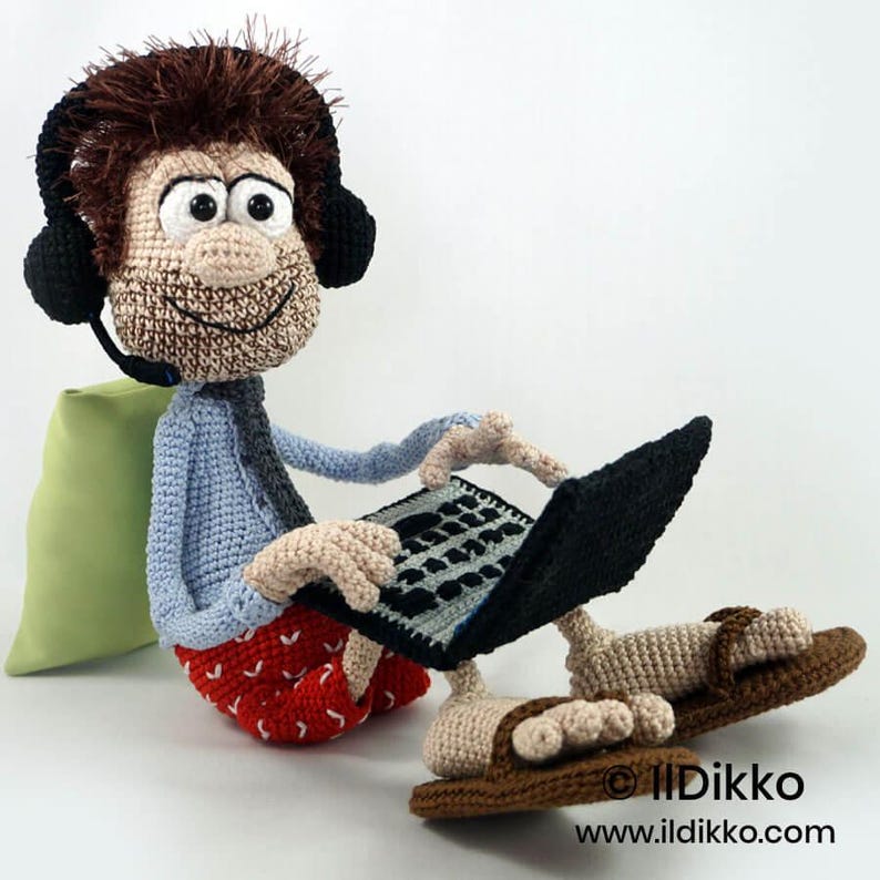 May include: A crocheted doll with brown hair, wearing black headphones with a microphone, a light blue shirt, and red patterned pants. The doll is sitting and using a black and gray crocheted laptop, with brown sandals on its feet.