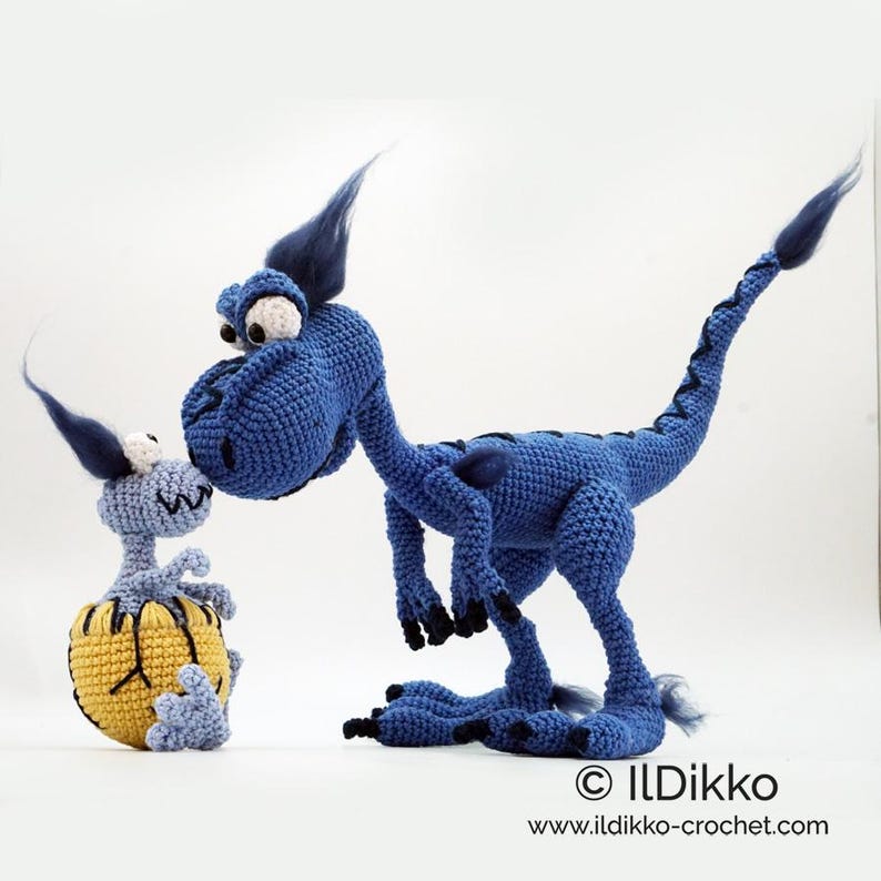 May include: A large blue crocheted dinosaur with dark blue tufts and black claws standing next to a smaller light blue crocheted baby dinosaur emerging from a yellow and black cracked egg. The text ILDikko www.ildikko-crochet.com is visible.