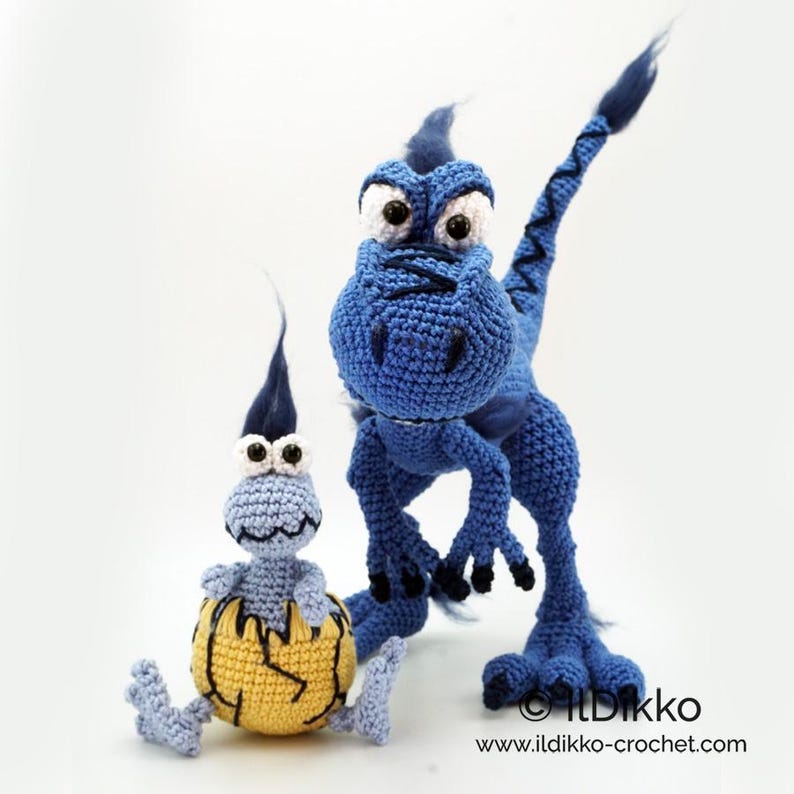 May include: Two crocheted dinosaur figures. One is a large blue standing dinosaur with dark blue accents and large eyes. The other is a smaller light blue dinosaur emerging from a yellow cracked egg. Text reads Ildikko www.ildikko-crochet.com.