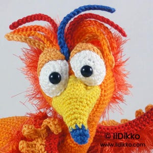 May include: A crocheted bird figure featuring bright orange and red yarn with a textured, fuzzy appearance. It has large white eyes with black pupils, a yellow beak with a blue tip, and blue and red yarn crests. Text reads: &copy; IlDikko www.ildikko.com