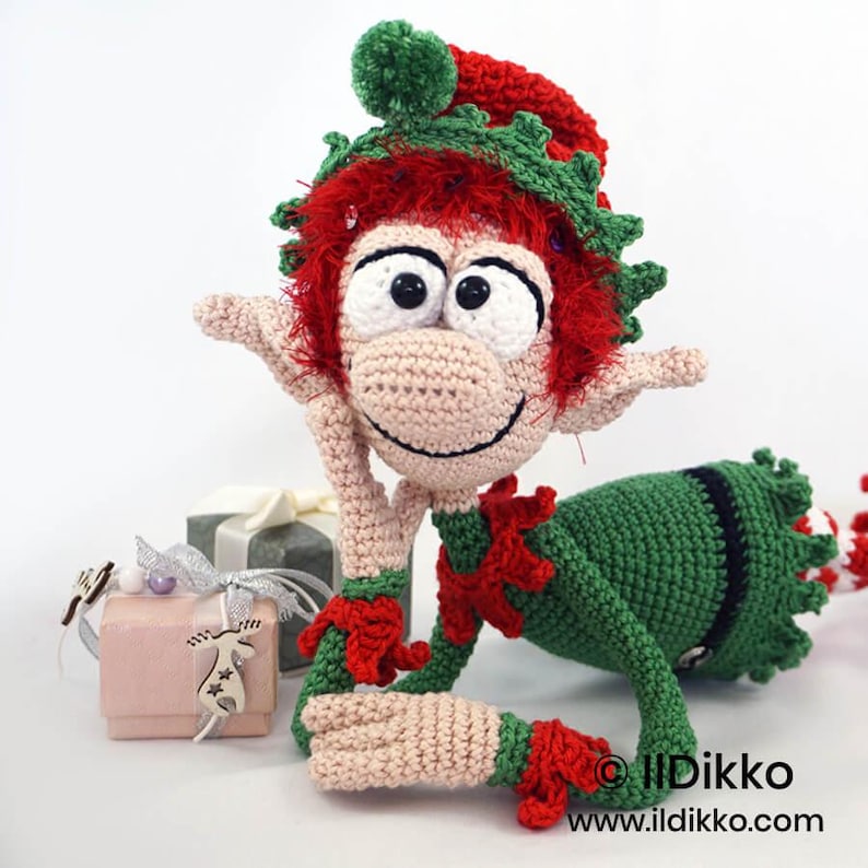 May include: A crocheted elf doll with red hair, a green hat with a pom-pom, and a green outfit with red accents. The doll has large white eyes with black pupils, a wide smile, and is positioned next to two small gift boxes with silver ribbons.