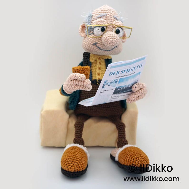 May include: A crocheted doll of an elderly man wearing glasses, holding a newspaper and a drink, seated on a small beige couch. The newspaper reads "DER SPIEGETTI". The doll is wearing a green jacket, yellow shirt, brown pants, and brown and tan slippers.