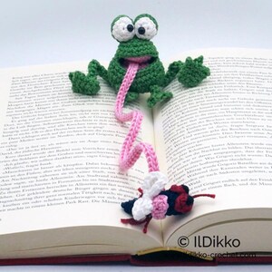 May include: A green crocheted frog bookmark with a pink tongue and a pink crocheted ribbon. The frog is sitting on an open book with a crocheted pink and white butterfly on the ribbon.
