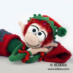 May include: A crocheted elf doll with beige skin, large white eyes, and bright red fuzzy hair. It wears a green and red hat with a pom-pom and green trim. The doll is positioned against red and green fabric. Text reads: &copy; IlDikko www.ildikko.com