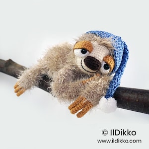 May include: A crocheted sloth toy with fuzzy light brown yarn, a dark brown nose, and black eyes. It wears a blue knit nightcap with a white pom-pom. The sloth is positioned on a dark brown branch. Text reads: © IlDikko www.ildikko.com.