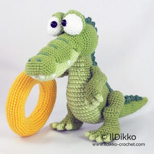 May include: A green crocheted crocodile toy with large white eyes and black pupils, holding a bright yellow crocheted ring in its mouth. The crocodile has a textured body, a long tail, and small claws. Text reads: &copy; Ildikko www.ildikko-crochet.com