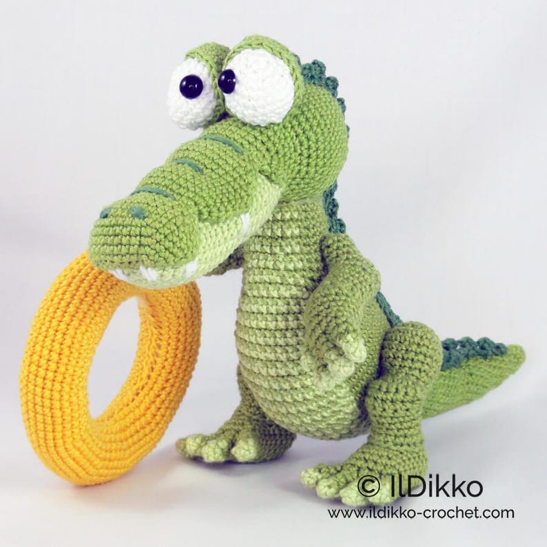 May include: A green crocheted crocodile toy with large white eyes and black pupils, holding a bright yellow crocheted ring in its mouth. The crocodile has a textured body, a long tail, and small claws. Text reads: &copy; Ildikko www.ildikko-crochet.com