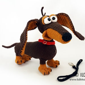 May include: A handmade crochet dachshund dog with brown and tan yarn. The dog has large, cartoonish eyes, a red collar, and a black leash. The dog is standing on a white surface.