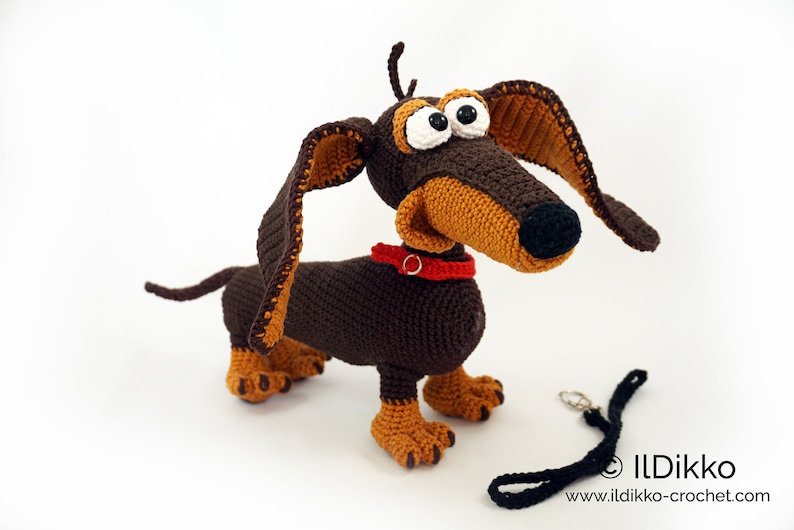 May include: A handmade crochet dachshund dog with brown and tan yarn. The dog has large, cartoonish eyes, a red collar, and a black leash. The dog is standing on a white surface.