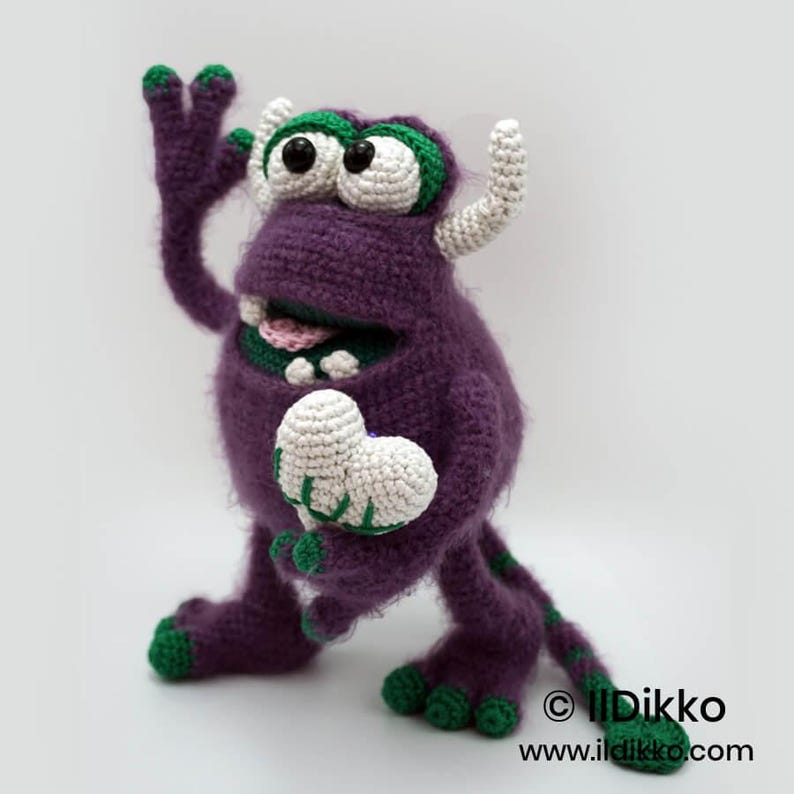 Puede incluir: Un mu&ntilde;eco monstruo de ganchillo morado con detalles verdes en los dedos de las manos y los pies, cuernos blancos y ojos grandes. El monstruo tiene la boca abierta mostrando dientes y una lengua rosa, y sostiene un coraz&oacute;n de ganchillo blanco con detalles verdes.