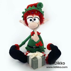 May include: A crocheted elf doll with red hair, large white eyes, and pointed ears, wearing a green tunic with a black belt and a red and green hat. The elf sits with one finger to its mouth, next to a small grey gift box with a white ribbon.