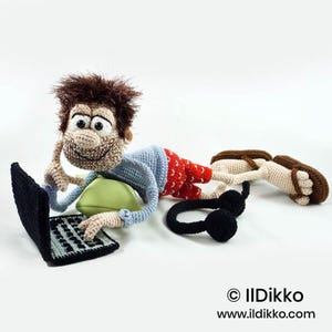 May include: A crocheted doll with brown hair, wearing a light blue long-sleeved shirt and red patterned shorts, lying on a light green pillow while using a black crocheted laptop. A pair of black crocheted headphones and brown sandals are nearby.