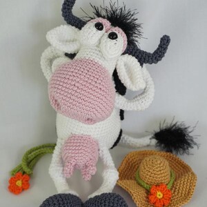 May include: A crocheted cow plush toy with black and white spots, pink accents, and a black and white tail. The cow is wearing a brown crocheted sun hat with a pink flower. The toy is sitting on a white surface.