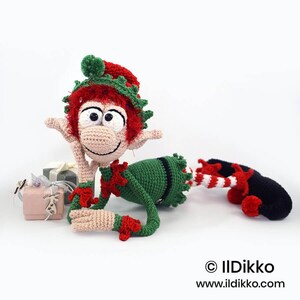 May include: A crocheted elf doll with red hair, a green and red hat, and a green outfit. The elf has large white eyes, a beige face, and pointed ears. It lies on its side next to two small gift boxes. Text reads: &copy; IlDikko www.ildikko.com
