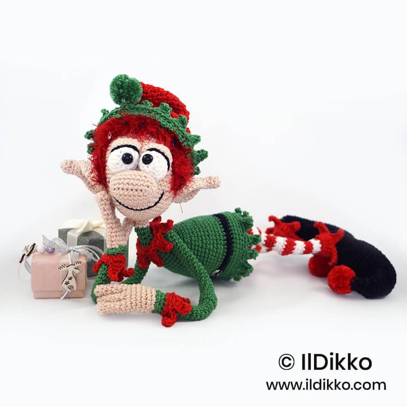 May include: A crocheted elf doll with red hair, a green and red hat, and a green outfit. The elf has large white eyes, a beige face, and pointed ears. It lies on its side next to two small gift boxes. Text reads: &copy; IlDikko www.ildikko.com