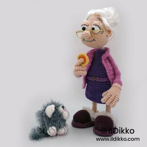 May include: A crocheted doll of an elderly woman wearing a purple dress and pink jacket, holding a yellow coin. A gray crocheted cat sits nearby. The doll has glasses and white hair. The doll is wearing purple shoes with white trim.