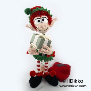 May include: A crocheted elf doll with red hair, a green hat with a pom-pom, and a green outfit. The elf has large eyes, pointed ears, and holds a small gift box with a white ribbon. It wears red and white striped leggings and black shoes.