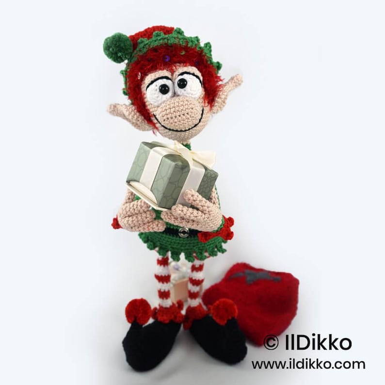 May include: A crocheted elf doll with red hair, a green hat with a pom-pom, and a green outfit. The elf has large eyes, pointed ears, and holds a small gift box with a white ribbon. It wears red and white striped leggings and black shoes.