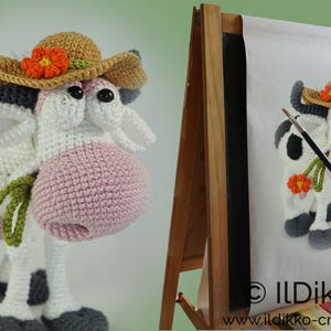 Peut inclure: Une vache en crochet portant un chapeau brun et orange avec une fleur. La vache tient une tige verte avec une fleur orange. La vache est debout sur une surface blanche.