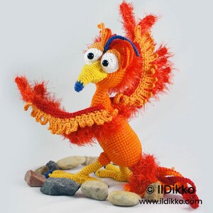 May include: A crocheted phoenix figurine featuring bright orange and yellow yarn with fuzzy red wing accents, a yellow beak with a blue tip, and large white eyes with black pupils. The creature has a blue crest and stands on yellow feet.
