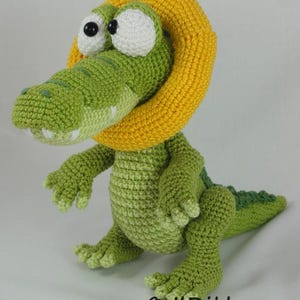 May include: A green crocheted crocodile toy featuring a large yellow ring around its neck, white eyes with black pupils, and a textured body. The text "&copy; ILDikko" and "www.ildikko-crochet.com" is printed in black at the bottom of the image.