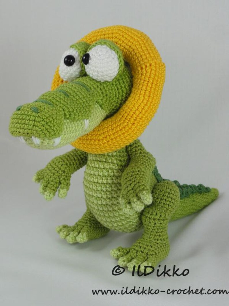 May include: A green crocheted crocodile toy featuring a large yellow ring around its neck, white eyes with black pupils, and a textured body. The text "&copy; ILDikko" and "www.ildikko-crochet.com" is printed in black at the bottom of the image.