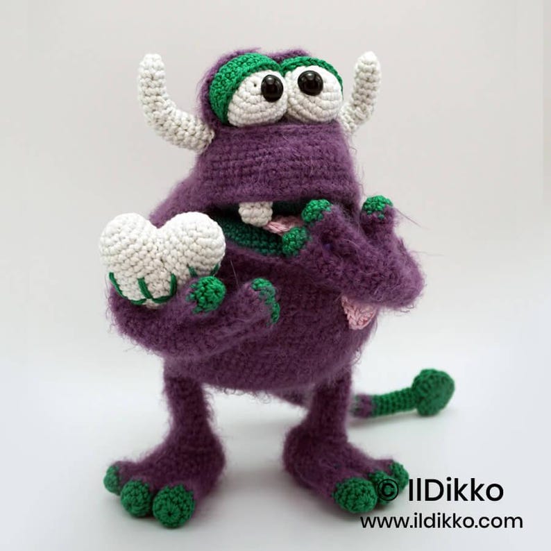 Puede incluir: Un monstruo de ganchillo morado con detalles verdes, dos cuernos blancos y grandes ojos blancos con pupilas negras. Sostiene un coraz&oacute;n de ganchillo blanco en una mano, tiene un solo diente y una cola verde. Texto: Ildikko ildikko.com.