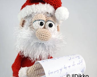 May include: A crocheted Santa Claus figure wearing a red hat with a white pom-pom and trim, featuring a white fuzzy beard, beige skin tone, and black eyes. The figure holds a long white scroll with blue cursive text and the text Ildikko ildikko.com.