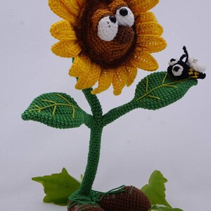May include: A crocheted sunflower with a brown face and large white eyes, wearing brown shoes. The sunflower has green leaves and a bee is perched on one of the leaves. The image includes the text "&copy; Ildikko www.ildikko-crochet.com".