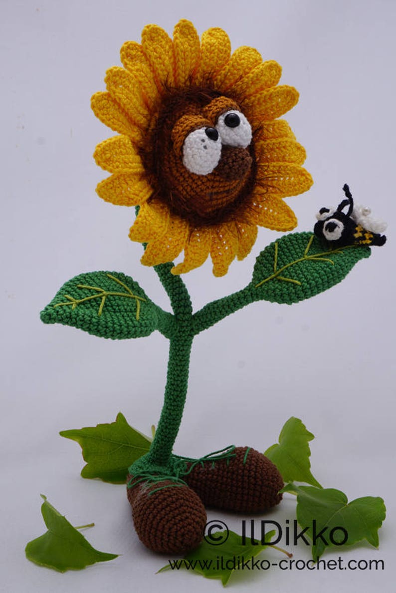 May include: A crocheted sunflower with a brown face and large white eyes, wearing brown shoes. The sunflower has green leaves and a bee is perched on one of the leaves. The image includes the text "&copy; Ildikko www.ildikko-crochet.com".