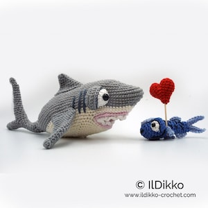 May include: A gray crocheted shark with a big toothy grin holds a red heart in front of a smaller blue crocheted fish.