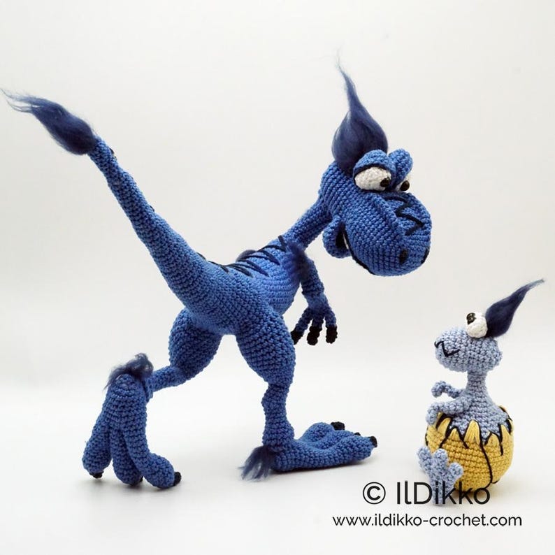 May include: A large blue crocheted dinosaur with dark blue tufts of hair and black claws standing next to a smaller light blue crocheted dinosaur emerging from a yellow cracked egg. The text ILDikko and www.ildikko-crochet.com is visible.