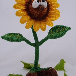 May include: A crocheted sunflower with a brown face, white eyes, and a black bee on top. The sunflower has a green stem and leaves, and brown feet.