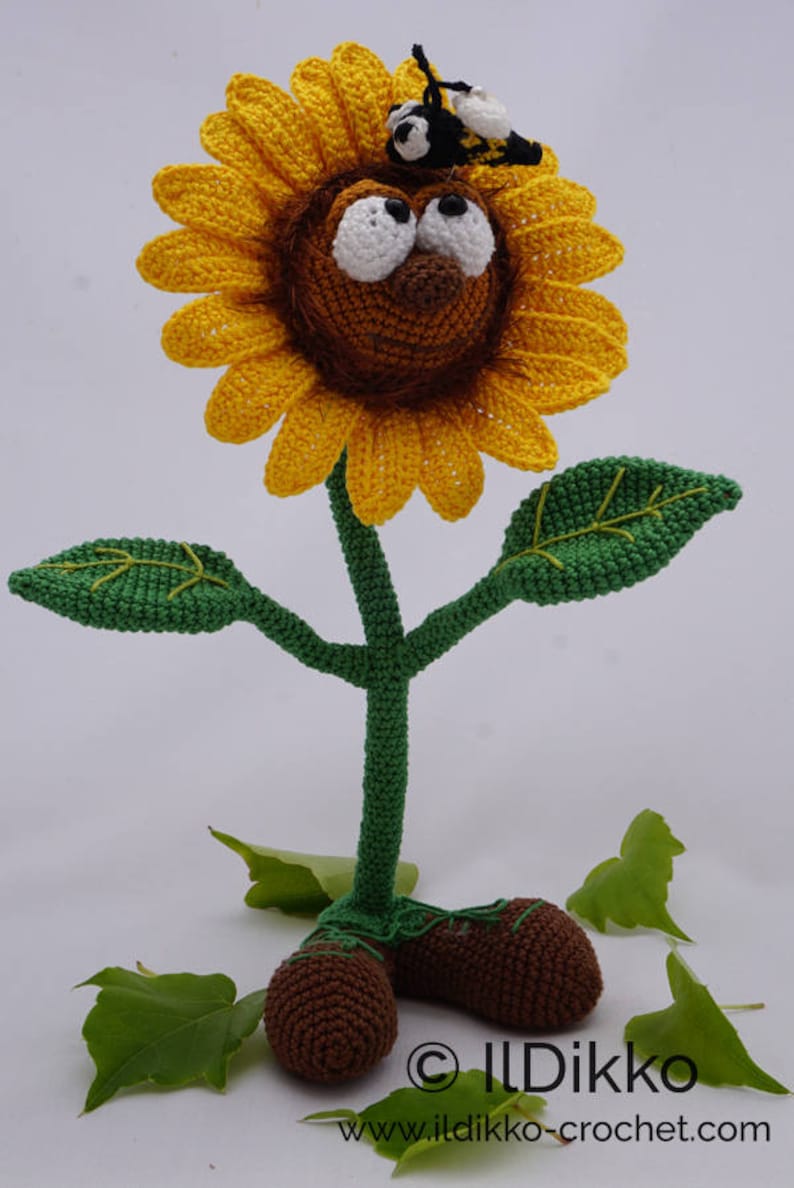 May include: A crocheted sunflower with a brown face, white eyes, and a black bee on top. The sunflower has a green stem and leaves, and brown feet.
