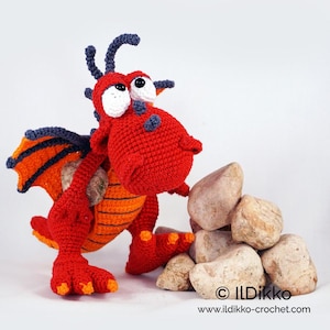 Puede incluir: Un dragón rojo de crochet con rayas naranjas y grandes ojos blancos. El dragón está sosteniendo una roca en sus patas delanteras y está de pie junto a una pila de rocas.