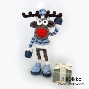 May include: A crocheted reindeer doll with a bright red nose, dark brown antlers, and a blue and white striped sweater, hat, and mittens. The reindeer wears blue boots with white trim and stands next to a small gift box with a cream ribbon.