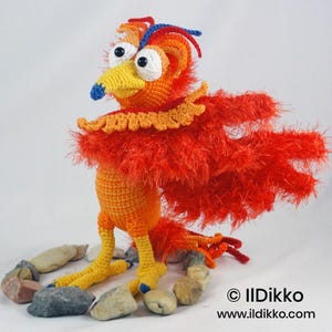 May include: A crocheted phoenix bird toy with a bright orange body, yellow legs, and long, shaggy red wings. It features large white eyes with black pupils, a yellow beak with a blue tip, and blue and red crest feathers. Text reads: &copy; IlDikko www.ildikko.com